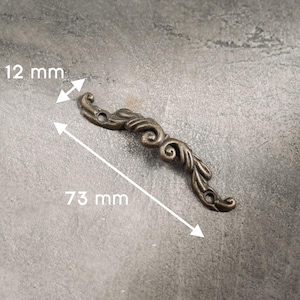 Puede incluir: Un adorno decorativo de mueble antiguo de color bronce, que mide 73 mm de largo y 12 mm de ancho. La pieza ornamentada presenta un diseño de volutas con una estética vintage, perfecta para añadir un toque de elegancia.