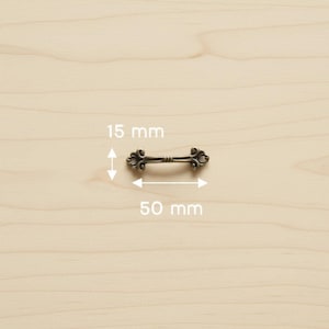 Op de afbeelding: Een antieke bronzen ladeknop met sierlijke krullen aan elk uiteinde. De knop is 5 cm lang en 1,5 cm hoog. De hardware wordt getoond op een licht houten oppervlak.