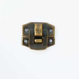 May include: A rectangular, antique brass-colored latch with a hinged closure. The latch has a rectangular body with rounded corners and two small holes on each side for mounting. The closure mechanism is a simple, rectangular clasp.