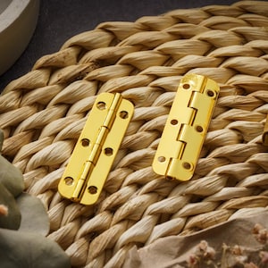 Könnte beinhalten: Zwei goldfarbene Metallscharniere mit dekorativem Design. Die Scharniere befinden sich auf einer geflochtenen Strohoberfläche.