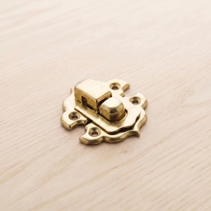 May include: A brass-colored, decorative clasp with a rounded, ornate design. The clasp features a rectangular latch and multiple screw holes for secure attachment. The hardware is set against a light wood grain background.