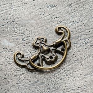 Puede incluir: Un tirador de cajón decorativo de latón adornado con un diseño curvo y abierto. El tirador tiene un patrón floral y un acabado liso y pulido.