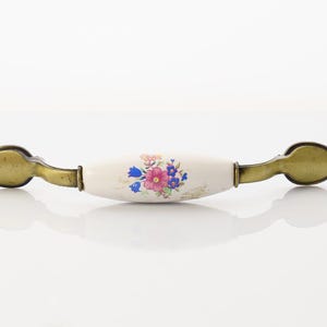 May include: A white ceramic cabinet knob with a floral design in shades of pink, blue, and green. The knob has a brass finish on the ends.
