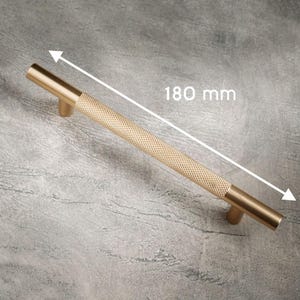 May include: A gold-colored cabinet pull with a diamond-patterned center and a total length of 180 mm (7 inches). The pull has a cylindrical shape with a smooth, solid end on each side.