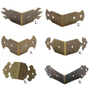 Pode incluir: Seis protetores de canto de metal em tom de latão para caixas ou móveis, cada um com uma forma e padrão decorativo distintos. Os itens estão numerados de 1 a 6, exibindo vários recortes, gravuras florais e furos para montagem.