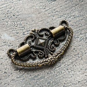 May include: A decorative brass drawer pull with an ornate design featuring a central square and intricate scrollwork. The pull has two cylindrical knobs.
