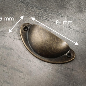 May include: A bronze-colored, half-moon shaped drawer pull with a distressed finish. The pull measures 33 mm wide and 81 mm long. It has two holes for mounting and is designed for furniture hardware.