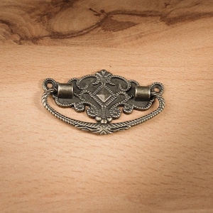 Può includere: Una maniglia per cassetti in stile antico con un design ornato. La maniglia in metallo ha una finitura scura e invecchiata e presenta un centro decorativo a forma di diamante. L'impugnatura è un design a corda curva. Lo sfondo è una superficie di legno chiaro.