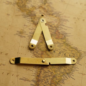 Puede incluir: Dos bisagras de metal dorado con una sola barra de metal dorado que las conecta. Las bisagras están abiertas y la barra está en el medio de las bisagras. Las bisagras y la barra están sobre un fondo marrón con un mapa impreso en él.