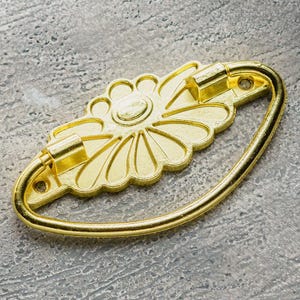 May include: Gold-toned metal drawer pull with a decorative flower design. The pull is shaped like a half-circle and has a smooth, polished finish.