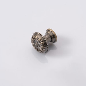 May include: A bronze-toned cabinet knob with a decorative, floral-like design on the face. The knob has a short, cylindrical stem and a circular base. The finish has an aged, antique appearance, suggesting a vintage or rustic style.