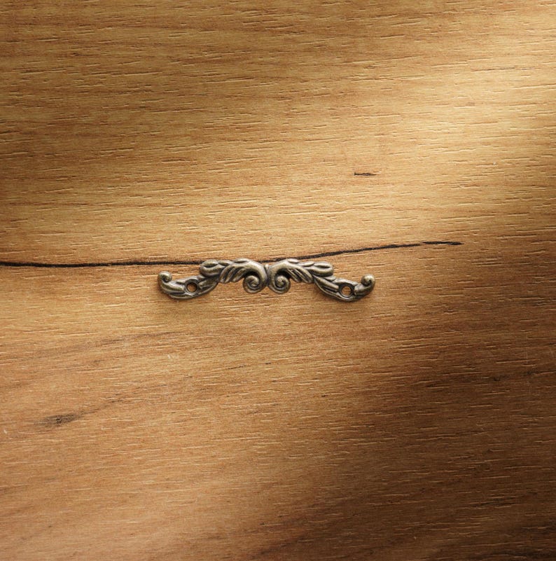 May include: A small, ornate, silver-colored metal furniture applique. The decorative piece features a symmetrical design with scrolling leaves and flourishes. The applique is set against a warm, light brown wooden surface.