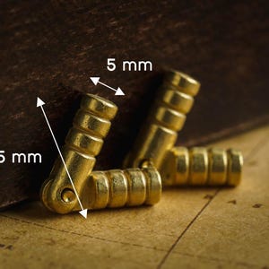 Op de afbeelding: Close-up van drie goudkleurige, L-vormige metalen scharnieren. Elk scharnier heeft een cilindrisch ontwerp met ribbels. De afmetingen zijn 2,5 cm bij 0,5 cm. De scharnieren worden weergegeven op een vintage kaart.