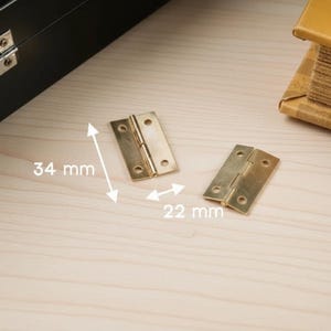 May include: Two brass-coloured hinges, each with three screw holes, are displayed on a light wood surface. The dimensions are 3.4 cm x 2.2 cm. The hinges are open and closed, showcasing their design.