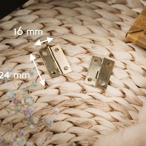 Puede incluir: Dos pequeñas bisagras de color latón, cada una con tres orificios para tornillos, se muestran sobre una superficie tejida. Una bisagra está abierta y la otra cerrada. Las dimensiones son 2,4 cm de alto y 1,6 cm de ancho.