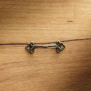 Puede incluir: Un tirador de cajón decorativo de color bronce antiguo con adornos de volutas en cada extremo. El tirador está centrado en una superficie de madera marrón claro, creando una estética vintage. El herraje está diseñado para muebles.
