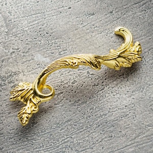 Op de afbeelding: Een gouden metalen ladegreep met een decoratief, sierlijk ontwerp dat lijkt op een gestileerd blad of tak.