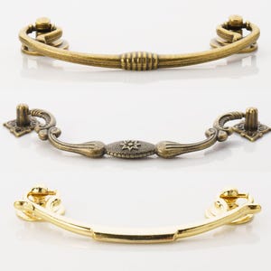 May include: Three antique-style cabinet door pulls. The top pull is a simple curved design in a brass finish. The middle pull is a more ornate design with a floral motif in a bronze finish. The bottom pull is a simple curved design in a gold finish.