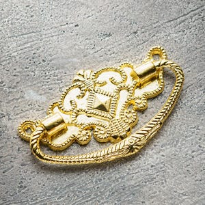 May include: A gold-toned metal drawer pull with an ornate design. The pull has a loop handle and a decorative centre piece.