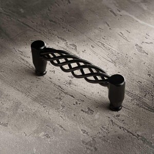 Puede incluir: Tirador de armario negro con un diseño decorativo de metal retorcido. El tirador mide aproximadamente 13 cm de largo y presenta un acabado mate. El tirador está diseñado para su uso en armarios, cajones o muebles.