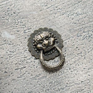 Puede incluir: Aldaba de puerta de bronce antiguo con diseño de cabeza de león. La aldaba presenta una cara detallada rodeada por un marco circular decorativo y un asa en forma de anillo. La ferretería está colocada sobre una superficie gris texturizada.