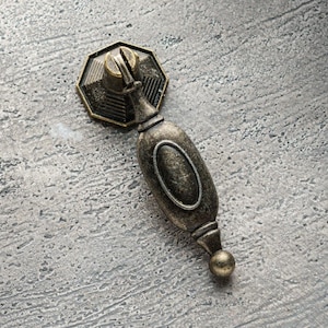 Puede incluir: Un tirador de armario de estilo vintage, adornado, de color bronce, con un diseño ovalado decorativo. El tirador tiene un pomo redondeado en la parte inferior.