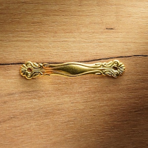 Puede incluir: Un tirador dorado y ornamentado con un diseño simétrico. El tirador presenta una sección central curvada y elementos florales decorativos en cada extremo. La ferretería está colocada sobre una superficie de madera.