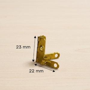 Puede incluir: Una escuadra de esquina dorada con dos orificios para tornillos. La escuadra mide 23 mm de alto y 22 mm de ancho. Los herrajes metálicos se muestran sobre un fondo claro de vetas de madera.
