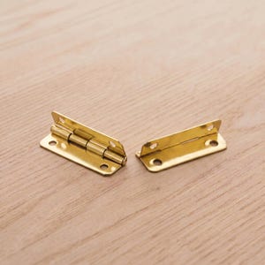 Op de afbeelding: Twee messingkleurige scharnieren, elk met drie schroefgaten, worden getoond op een licht houten oppervlak. De scharnieren zijn open en onthullen de cilindrische pen die de twee rechthoekige platen verbindt. Beslag voor meubels of kleine dozen.