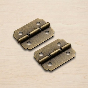 Puede incluir: Dos bisagras de color latón antiguo. Cada bisagra tiene una placa rectangular con bordes redondeados y tres agujeros circulares. Un pasador cilíndrico conecta las dos placas. Para muebles o proyectos decorativos.