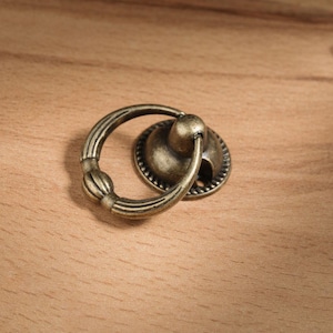 May include: Antique brass-toned drawer pull with a ring handle and a decorative backplate. The ring handle features a ribbed design with a central, decorative element. The backplate has a circular shape with a beaded edge. The hardware is set against a light brown wooden surface.
