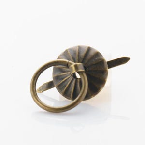 Puede incluir: Un tirador de anillo de metal de color bronce con una parte superior decorativa y estriada. El tirador de anillo está diseñado para sujetarse a una caja, bolsa u otro objeto.