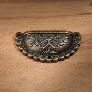 May include: Antique bronze drawer pull with a decorative dragon design. The pull has a curved shape with a scalloped edge and two mounting holes. The dragon is centered on the pull.