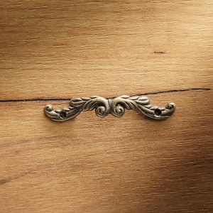 Puede incluir: Un tirador decorativo de estilo antiguo con un diseño ornamental y simétrico. El tirador de metal es de color plateado apagado y está montado sobre una superficie de madera con veta visible.