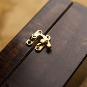 May include: A close-up shot of a dark brown wooden box with a gold-coloured clasp. The box has a smooth, polished finish, and the clasp is securely fastened. The wood grain is visible, adding texture to the surface. The box appears to be new.