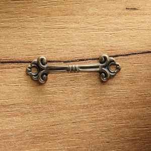 Op de afbeelding: Een kleine, sierlijke metalen ladetrekker in antieke stijl met een donkere, verouderde afwerking. De trekker is een horizontale staaf met decoratief krulwerk aan elk uiteinde. Het is gecentreerd op een houten oppervlak. De trekker is ongeveer 7,6 cm lang.
