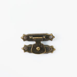 May include: A small, antique-style metal clasp in a bronze finish. The clasp is composed of two ornate, rectangular pieces with decorative edges and a central hinge. It is designed to secure a box or similar item.