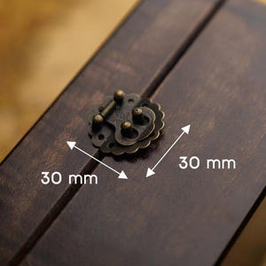 Puede incluir: Primer plano de una caja de madera con un cierre decorativo de latón antiguo. La caja y el cierre son de color marrón oscuro. El cierre mide 30 mm de largo.
