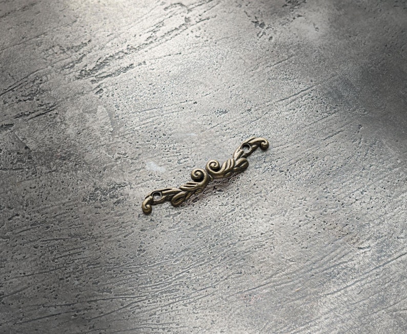 May include: A small, ornate, antique brass-colored metal decorative element. The piece features a symmetrical design with scrolling flourishes and leaf-like details, suitable for craft projects or furniture embellishment.