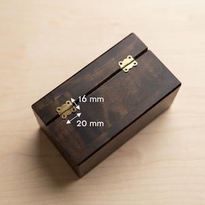 Puede incluir: Una caja de madera teñida oscura con tapa con bisagras y cierres de color latón. La caja es rectangular y parece estar vacía. Los cierres miden 20 mm de ancho y 16 mm de alto. La caja está sobre una superficie de madera clara.