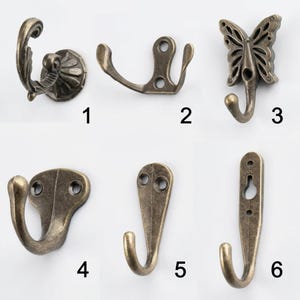 Op de afbeelding: Een collectie van zes metalen wandhaken met een antieke bronsafwerking, genummerd van één tot zes. Elke haak heeft een eigen vorm, waaronder sierlijk krulwerk, een vlinderontwerp en diverse montageplaten met schroefgaten.