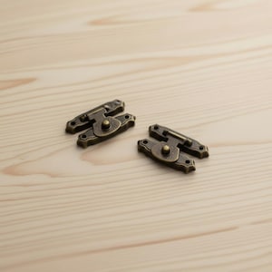 May include: Two antique bronze-coloured latches with a decorative design. Each latch has a central circular element and a hinged bar. The latches are designed for use on small boxes or chests.