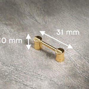 Puede incluir: Un pequeño tirador de armario dorado con una barra que conecta dos extremos cilíndricos. El tirador mide 1 cm de alto y 3,1 cm de largo. La ferretería está sobre un fondo gris moteado.