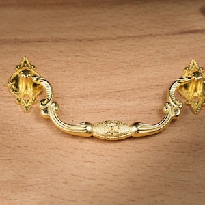 May include: A gold-colored drawer pull with an ornate design. The pull features a curved handle with decorative detailing and diamond-shaped mounting plates. The hardware is likely for furniture restoration or a decorative project.