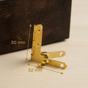 Op de afbeelding: Een goudkleurig metalen hoekscharnier, 30 mm hoog en 32 mm breed, wordt getoond tegen een houten achtergrond. Het scharnier is in een hoek van 90 graden ontworpen en heeft meerdere schroefgaten. De afbeelding toont de afmetingen van het scharnier.