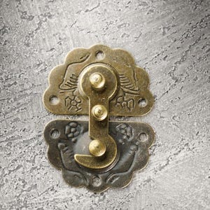 May include: Ornate brass-toned clasp with decorative floral and bird motifs. The clasp is composed of two pieces, a top and bottom, connected by a pivoting mechanism. The background is a textured, gray surface.