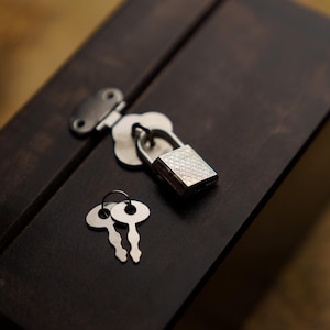 Puede incluir: Una caja de madera marrón oscuro con un candado plateado y dos llaves a juego. El candado está cerrado y las llaves están colocadas sobre la caja. La caja tiene forma rectangular y una tapa con bisagras.
