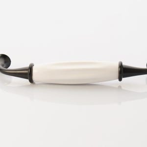 May include: A cabinet pull with a white ceramic center and black metal ends. The handle has a curved design with a rounded knob on each end. The hardware is designed for furniture and home decor.