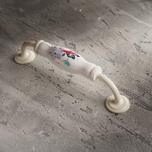 May include: A white ceramic cabinet pull with a floral design in pink, purple, and green. The handle has a curved shape and is mounted on a gray surface.