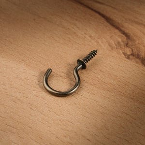 Könnte beinhalten: Nahaufnahme eines einzelnen antiken, bronzefarbenen Schraubhaken. Der Haken ist gebogen und hat ein Gewinde zum Einschrauben. Ideal zum Aufhängen von Gegenständen, ist diese Hardware eine praktische Ergänzung für jedes Zuhause oder jede Werkstatt.